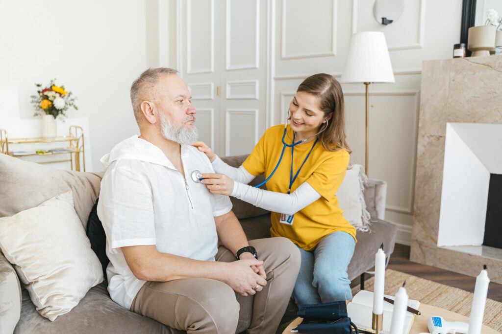 pexels-photo-7345465-7345465 A nurse uses a stethoscope for a home check-up on a senior adult in a cozy living room.